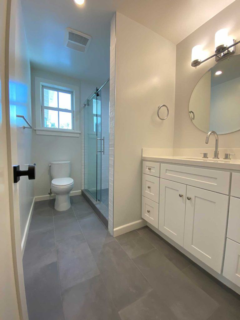 bathroom with white vanity and circular mirror with toilet and glass door shower in the back